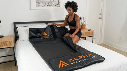 Woman using an Alpha Recovery Lab sauna blanket on a bed in a bright bedroom recovery setting.